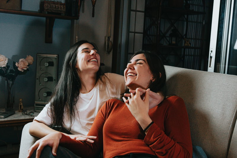 Photo of two women lounging on a sofa. They're snuggling and laughing