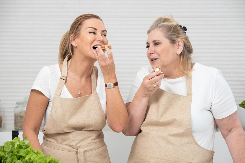 Two middle-aged white women wearing aprons, standing in the kitchen, sharing dessert, and laughing