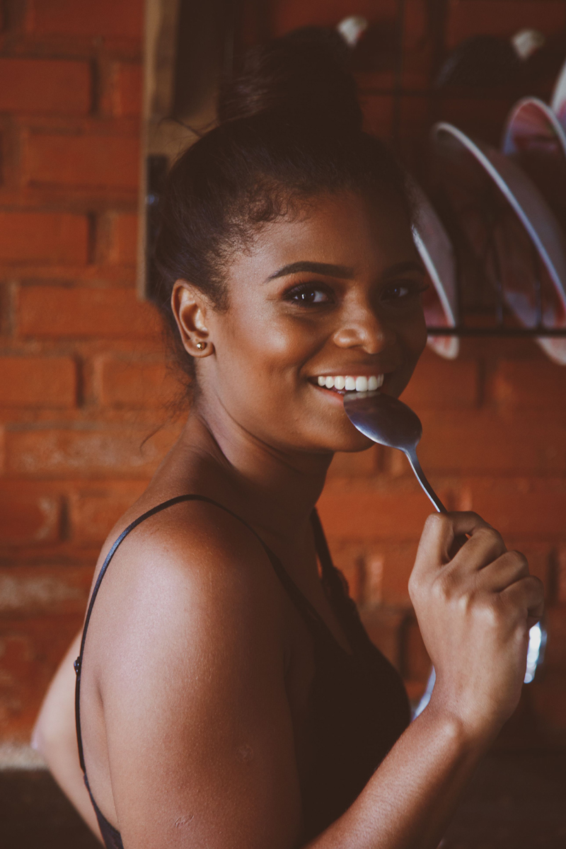 Portrait photo of young Black woman, looking towards her right, smiling at the camera, and holding a spoon to her mouth
